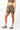 A model wearing high-waisted athletic biker shorts in a taupe brown color, paired with a matching sports bra, beige socks, and trail running shoes against a light gray studio background.