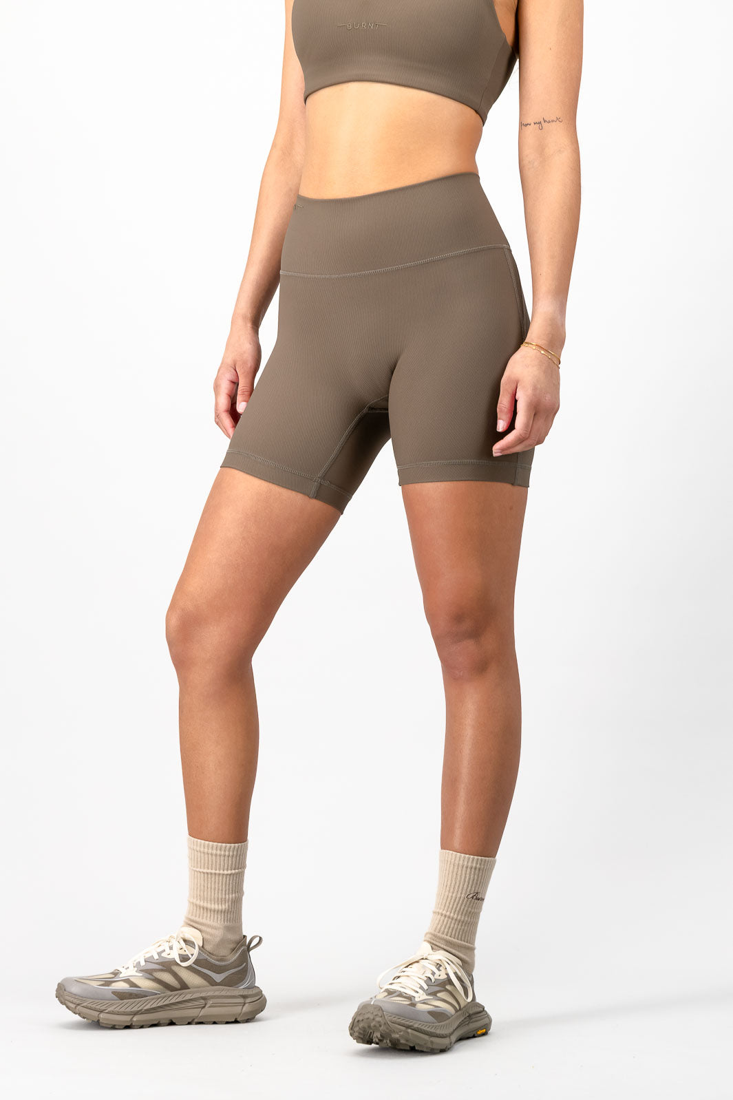 A model wearing high-waisted athletic biker shorts in a taupe brown color, paired with a matching sports bra, beige socks, and trail running shoes against a light gray studio background.