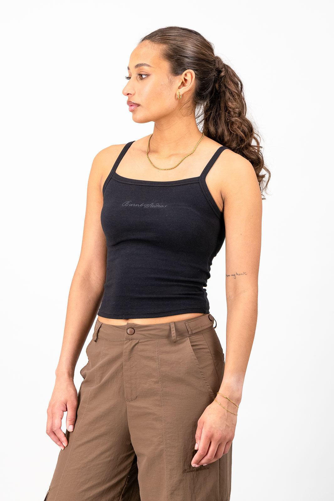 A woman wearing a black square-neck tank top with thin straps and a small white script logo on the chest, paired with brown pants, photographed against a plain white studio background.